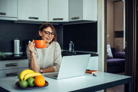 Happy young female blogger wearing glasses in a home kitchen with laptop broadcasting live. Communication with subscribers, remote work, social distance.の写真素材