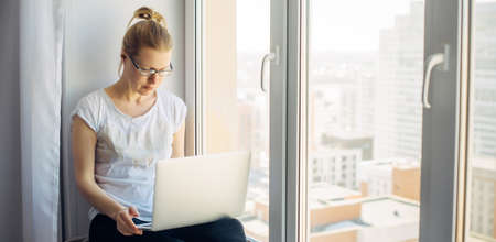 Young blonde woman wearing glasses in white t-shirt working on laptop at home, sitting on the windowsill. Freelancer and remote business. Window background. Copy space.の写真素材