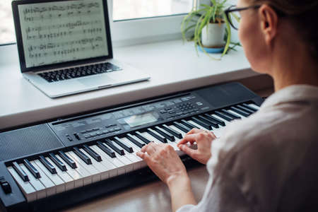 Back view young woman playing a synthesizer, reading notes on a laptop screen, close up. Independent learning to play the piano at home. Passion for music, hobbies, leisure, self-development.の写真素材