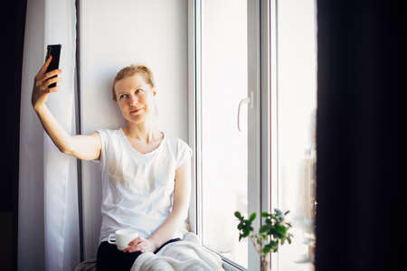 Young adult woman with glasses takes a selfie sitting on the windowsill at home, gesturing with her hand. Female smiles while chatting, looking at smartphone.の写真素材