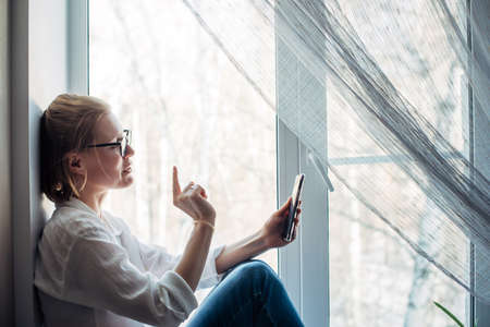 Happy woman in glasses and white shirt sits on the windowsill, holding smartphone in her hand. Cute blonde smiles at phone camera, makes hand gesture, close-up. Chat online.の写真素材