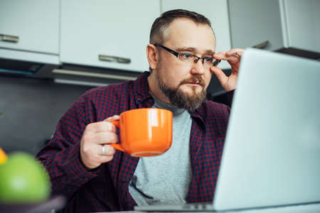 Stylish intelligent man with a beard in modern minimalist kitchen interior. Businessman in a home shirt having breakfast and studying the news looking at laptop.の写真素材