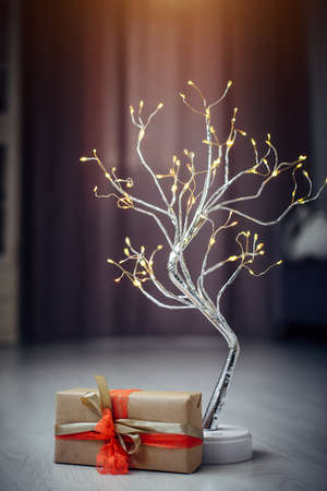 Beautiful silver tree with lighting, and gift box decorated with ribbons and bow in stylish home interior. New Year's still life - metal garland and box of craft paper on a blurry background.の写真素材
