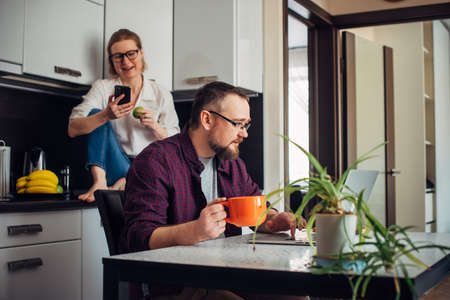 Adult married couple in home kitchen interior. Husband with beard sitting at table, looking at...