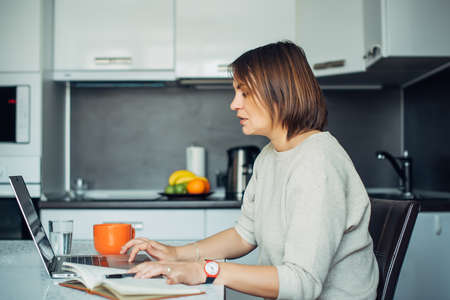 Young pretty brunette with laptop sitting at a table in the kitchen. Female writer works at home and drinks coffee. Freelance, remote work, creativity.の写真素材