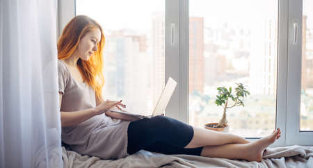 Young attractive brunette sitting on the windowsill, with laptop on her lap, typing on keyboard. Beautiful long-haired woman works at home, in city apartment.の写真素材