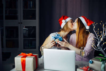 Loving female couple giving New Year's gifts at home interior. Two young women in Santa Claus hats sitting at table with laptop and wishing Merry Christmas. LGBT family concept.の写真素材