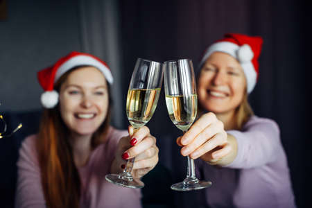 Christmas toast with glasses of champagne in hands. Blurred background: two girls in Santa Claus hats, clink glasses and wish Happy New Year. Love, friendship, family.の写真素材