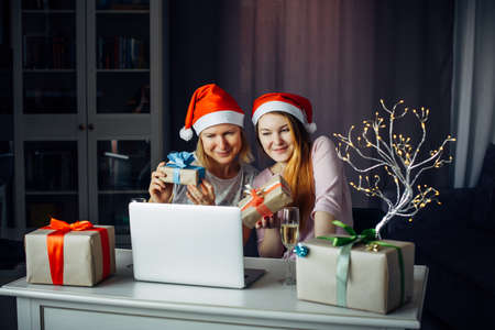Two pretty women in Santa Claus hats sitting at table with laptop among garlands and gifts and wish a Merry Christmas. Communication with friends and family via the Internet on New Year's Eve 2022.の写真素材