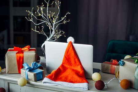 New Year's still life in home interior, close-up. Santa Claus hat hanging from laptop, gift boxes and festive garlands. Christmas card with copy space.の写真素材
