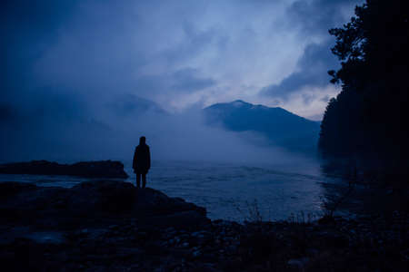 Human silhouette in a blue haze on the background of mountains and river. Thick fog in the evening twilight. Mysterious atmosphere. Concept of solitude, reflection, meditation.の写真素材