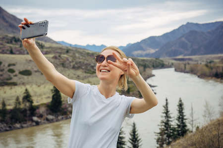 Young blonde girl in sunglasses and white T-shirt takes a selfie on her smartphone on the background of river and rocky mountains. Happy female tourist smiles at mobile phone camera.の写真素材
