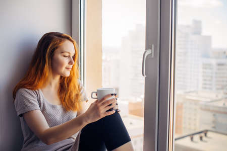 Beautiful young woman sitting on windowsill overlooking the metropolis, drinking tea or coffee from cup. Female portrait on the background of window in city apartment. Peace of mind and mental health.の写真素材