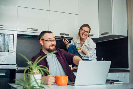 Happy forty-year-old married couple spending time together in home kitchen. Man looks at laptop and points at the monitor, while a woman with glasses sits behind and laughs. Focus on wife.の写真素材