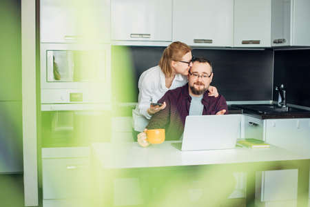 Happy adult married couple using laptop in cozy kitchen interior. Woman in glasses embracing her husband. In the foreground leaves of houseplant, blurred focus. Family mutual understanding concept.の写真素材