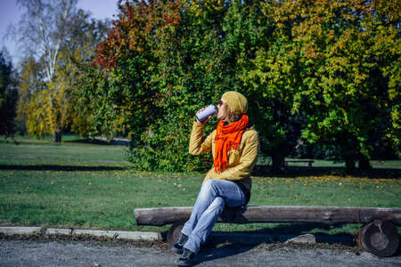 Pretty european woman in yellow jacket and red scarf sitting on wooden bench in a deserted park and drinking coffee. Enjoying solitude and sunny autumn weather. Blurred plant background, copy space.の写真素材