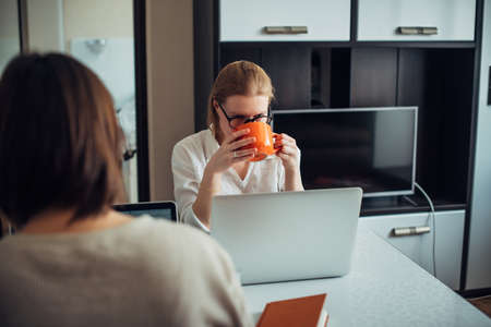 Two young adult girls sitting at table facing each other, working on laptops in the home interior. Focus on face of blonde with glasses. Freelance, coworking, remote work, self-employment.の写真素材
