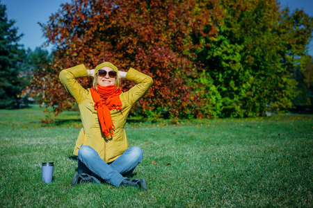 Carefree hipster woman with raised hands sitting on green lawn enjoying good weather. Outdoor coffee...
