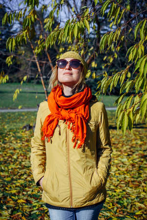 Attractive girl in sunglasses and red scarf smiling on the background of yellow foliage in autumn park. Young positive woman enjoying walk on fall sunny day.の写真素材