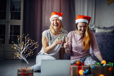 Two happy excited young woman in Xmas Santa hats toasting with fizzy wine at laptop webcam. Female love couple making New Year video call from home.の写真素材