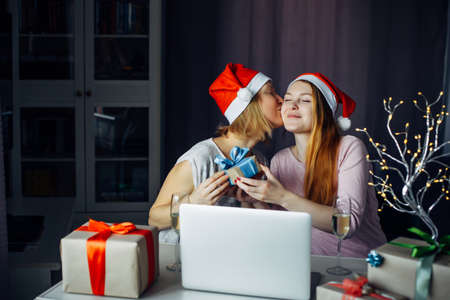 Loving female couple giving New Year's gifts at home interior. Two young women in Santa Claus hats sitting at table with laptop and wishing Merry Christmas. LGBT family concept.の写真素材