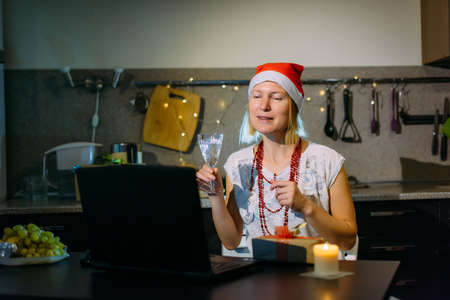 Joyful middle-aged woman in red Santa's hat looks at laptop and holds a glass of champagne, sitting at home kitchen in the light of garland. Remote New Year greetings from friends and family.の写真素材