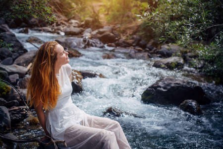 Dreaming romantic woman in white dress sitting on mountain river bank among green trees. Young girl raising her face to the sun on nature background. Harmony happy lifestyle concept.の写真素材
