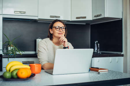 Happy young female blogger wearing glasses in a home kitchen with laptop broadcasting live. Communication with subscribers, remote work, social distance.の写真素材