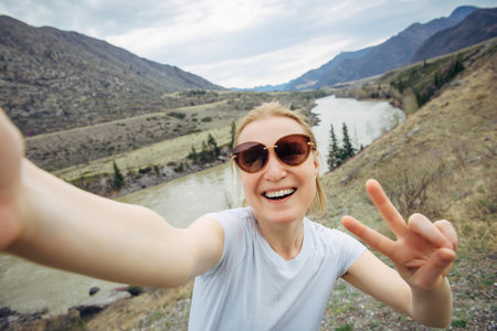 Happy young adult woman in sunglasses and white T-shirt takes a selfie, laughing into a smartphone camera against the mountain river landscape. Travel, blogging, active lifestyle.の写真素材