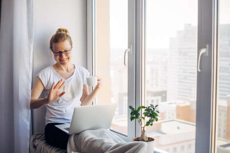 Happy smiling woman wearing glasses sitting on windowsill at city apartment, resting, looking at webcam on laptop, talking on video chat with her friends or family.の写真素材