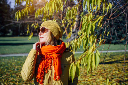Portrait of attractive girl in sunglasses and red scarf on the background of yellow foliage in autumn park. Young woman enjoying walk on fall sunny day.の写真素材