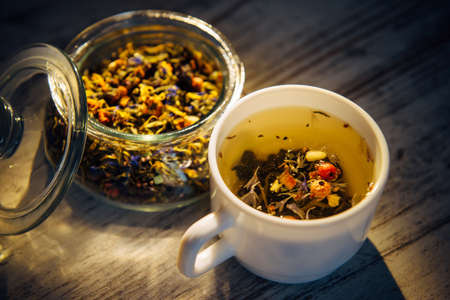 Beautiful blended tea made of herbs, berries and nuts in white mug and transparent glass bowl on vintage wooden background, close-up. Brewing tea in the evening light. Healthy lifestyle, detox.の写真素材