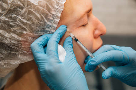 Adult woman with wrinkles around her eyes in process rejuvenation injections of hyaluronic acid filler, close up. Cosmetologist injects botulinum toxin to smooth female face skin.の写真素材