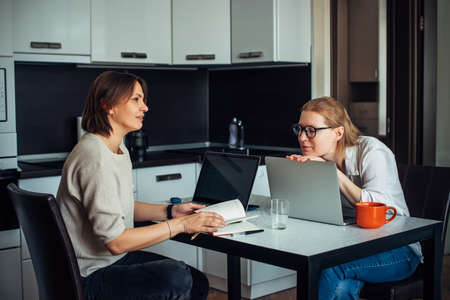 Happy emotional women discuss creative work issues sitting at kitchen table with laptops and notebooks. Coworking, freelance, startup, remote work.の写真素材