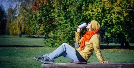 Pretty european woman in yellow jacket and red scarf sitting on wooden bench in a deserted park and drinking coffee. Enjoying solitude and sunny autumn weather. Blurred plant background, copy space.の写真素材