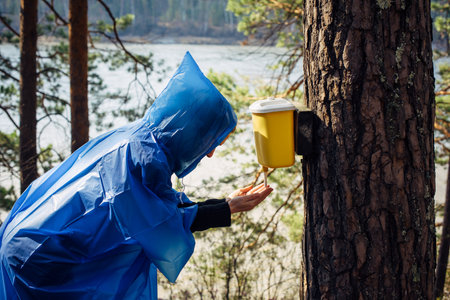 Woman in blue raincoat washes her hands in wash basin hanging on tree. Morning after rain at tourist camp in the forest by the river. Girl's face hidden by hood. Hiking lifestyle.の写真素材