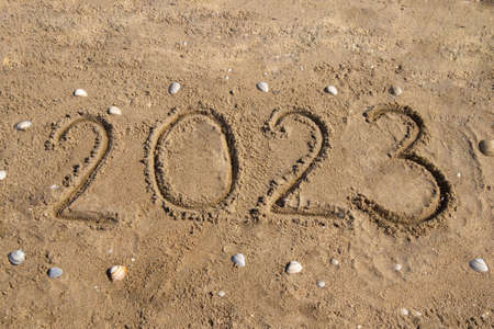 The numbers 2023 are handwritten on the sand, on the seashore, around the seashell.