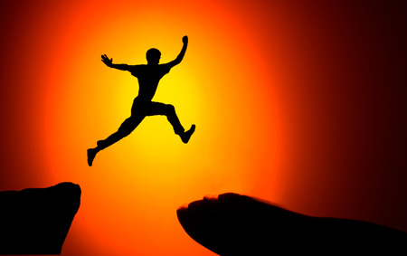 Man jumping across the gap from one rock to cling to the other. Man jumping over rocks with gap on sunset fiery background. Element of designの写真素材