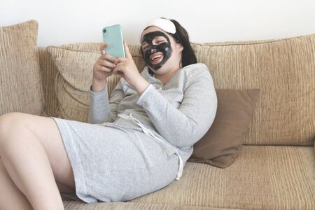 young woman at home with charcoal mask on her face on the couch holding a smartfone calling her friends and having funの写真素材
