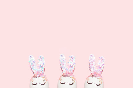 cute Easter bunny shape eggs with eyes and shiny glitter and confetti ears on a pastel pink...
