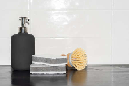 stylish cleaning supplies concept black soap dispenser, white cleaning sponges and a wooden bamboo brush on a black counyter top and white tilesの写真素材