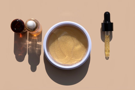 close up of pipette dropper with face serum, eye patches and serum bottles on a beige background...