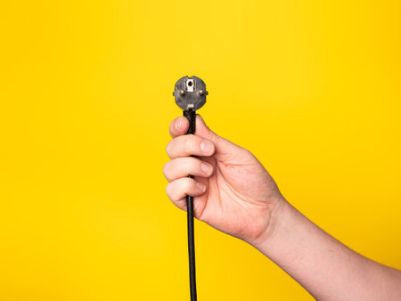 A hand holds a plug to which a black cable is attached. No face, yellow background.の写真素材