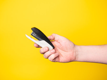 A hand holding a black and white stapler on a yellow background.