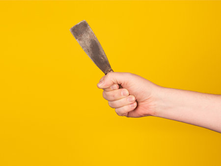 A man's hand holds a metal spatula on a yellow background
