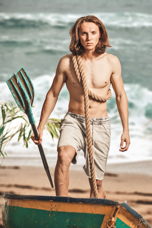 sexy man half naked, caucasian, long-haired with oar in boat on sand coast of desert islandの写真素材