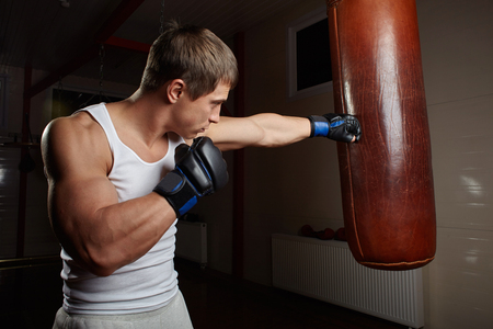 young muscular male boxer punch punching bag in gym interiorの写真素材