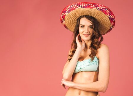 young happy beautiful female in bikini and straw hat looking at camera on pink backgroundの写真素材