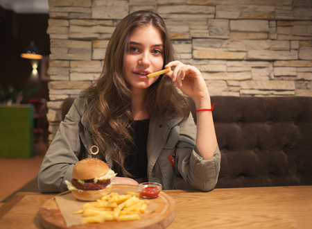 young lovely woman sitting in cafe with burger and chips, eating french friesの写真素材