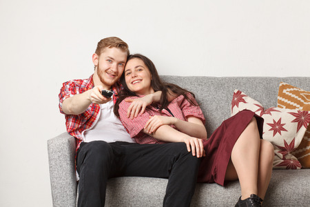 young couple, guy with pretty girl, watching TV on sofa and switch channel with remote controllerの写真素材